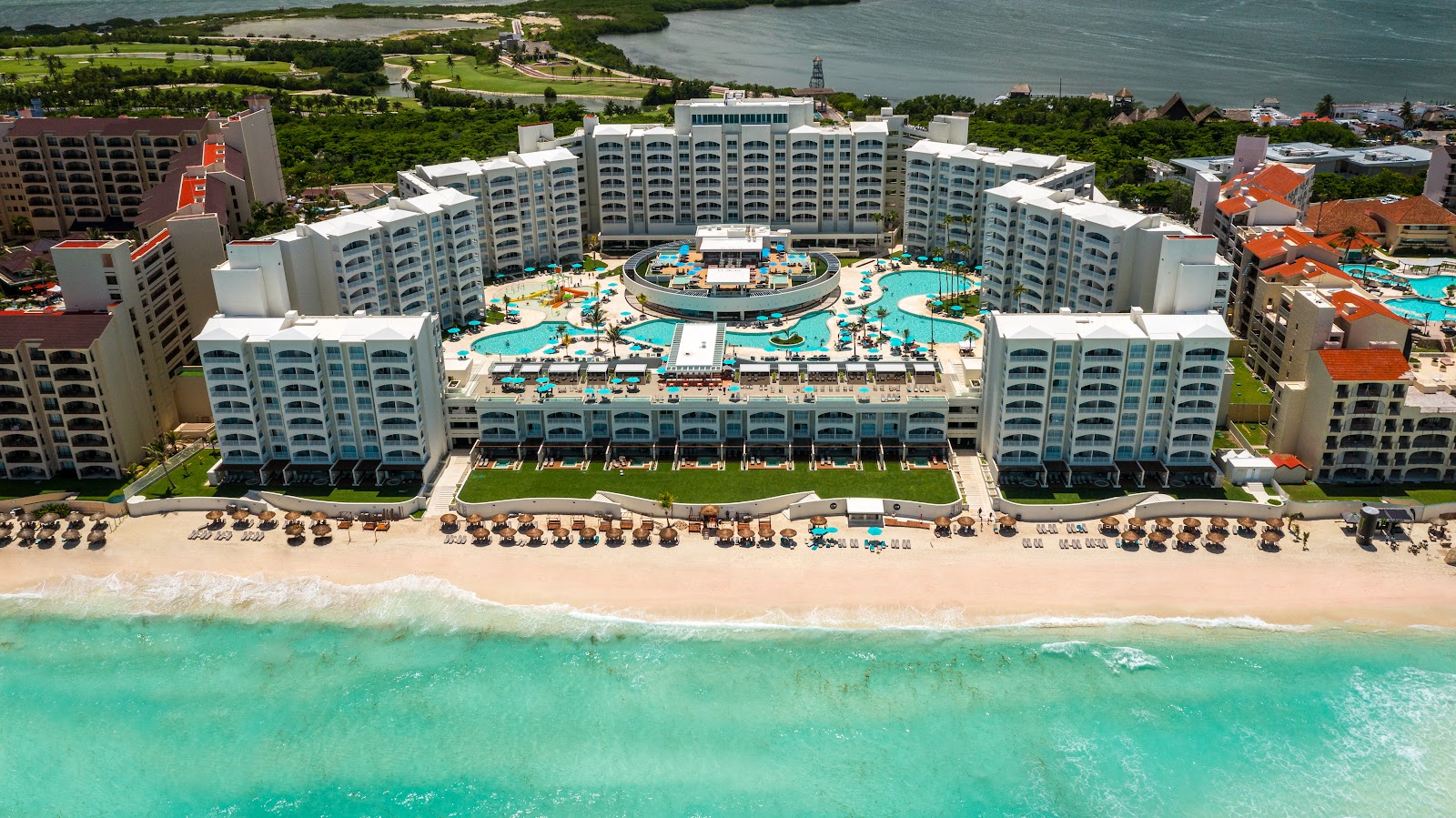 Aerial view of Hilton Cancun Mar Caribe with turquoise ocean, sandy beach, loungers, and multiple pools surrounded by white high-rise buildings.