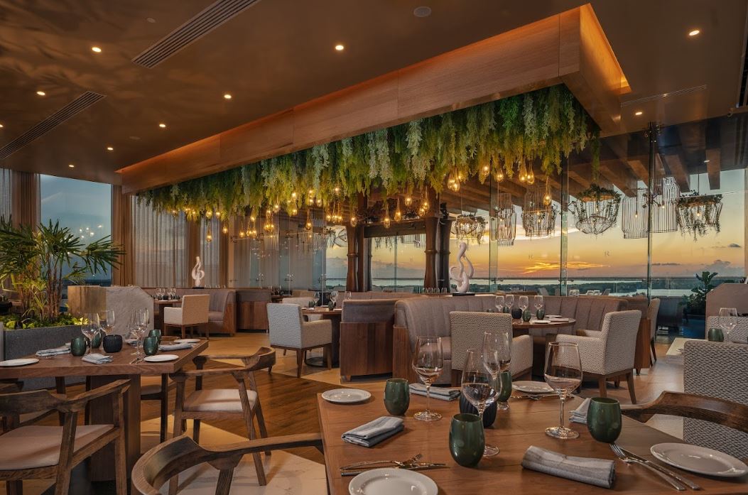 Hilton Cancun Mar Caribe restaurant with wooden tables, tan chairs, hanging greenery with pendant lights, and ocean sunset view through floor-to-ceiling windows.