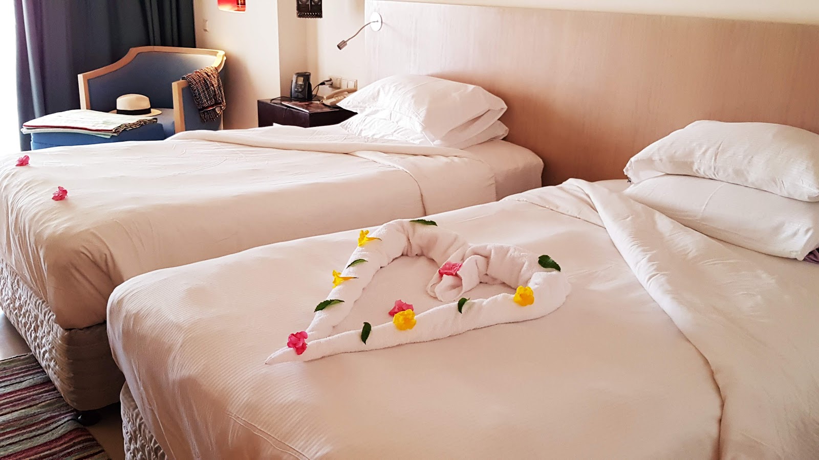 Resort guestroom with three beds decorated with pink and yellow flowers, bedside tables, chair, and purple curtains.