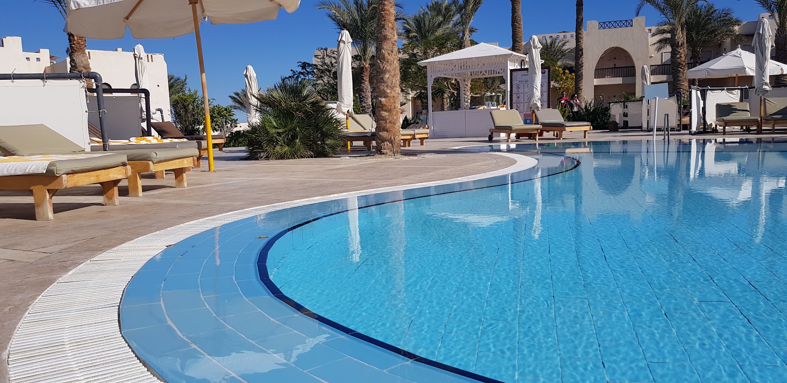 Curved pool with pale deck, wooden loungers under umbrellas, palm trees, and whitewashed resort buildings under clear blue sky.