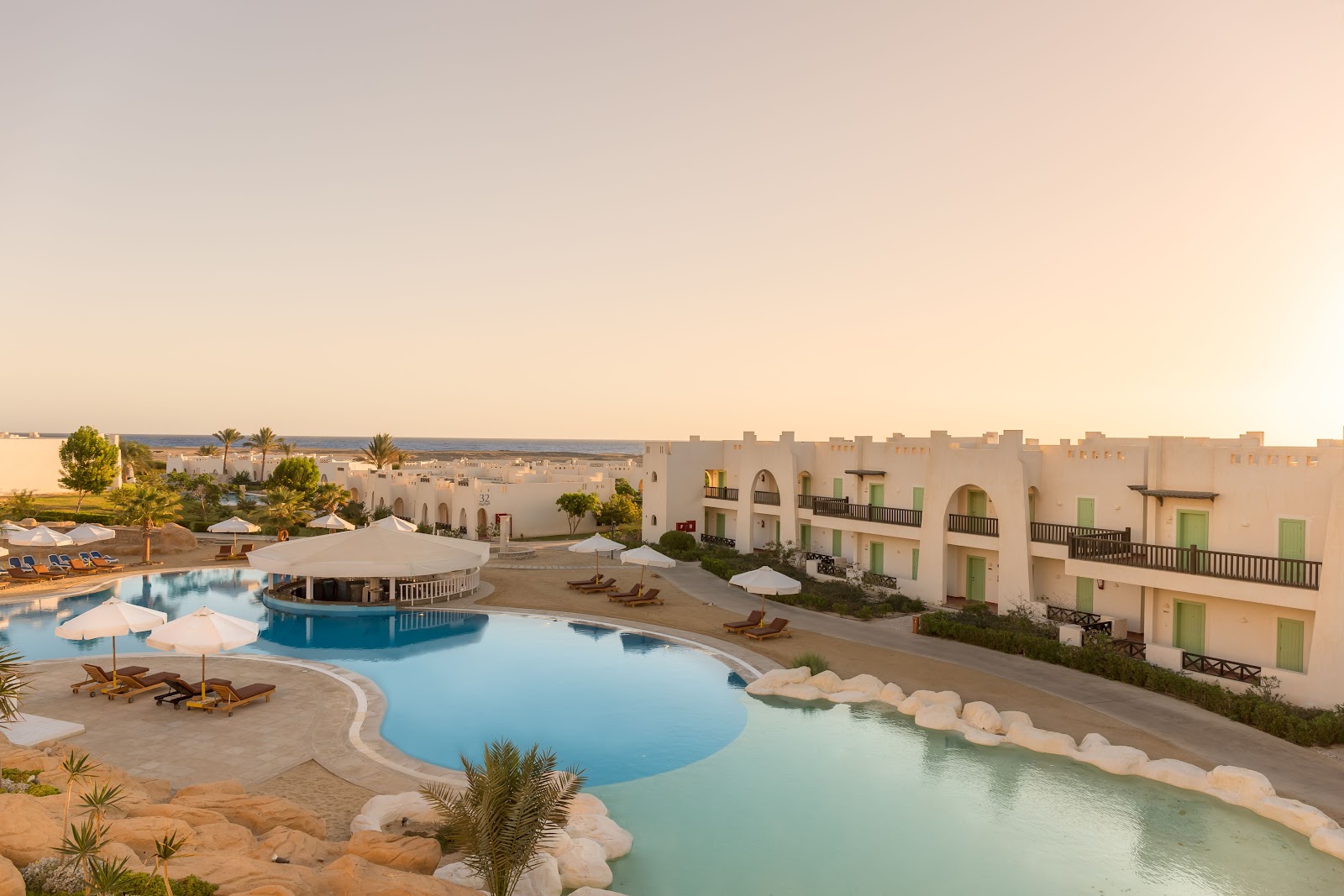 White resort buildings with green shutters overlook curved pools with loungers and umbrellas at sunset in the Egyptian desert.