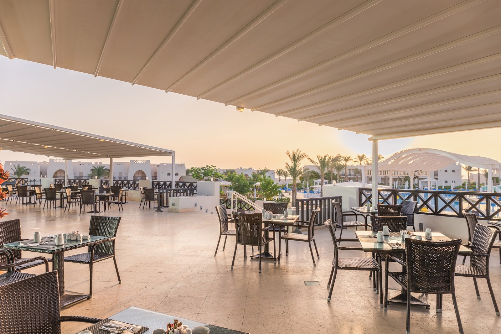 Shaded terrace with dark wicker chairs and tables overlooking white resort buildings, palm trees, and a pool at Hilton Marsa Alam Nubian Resort.