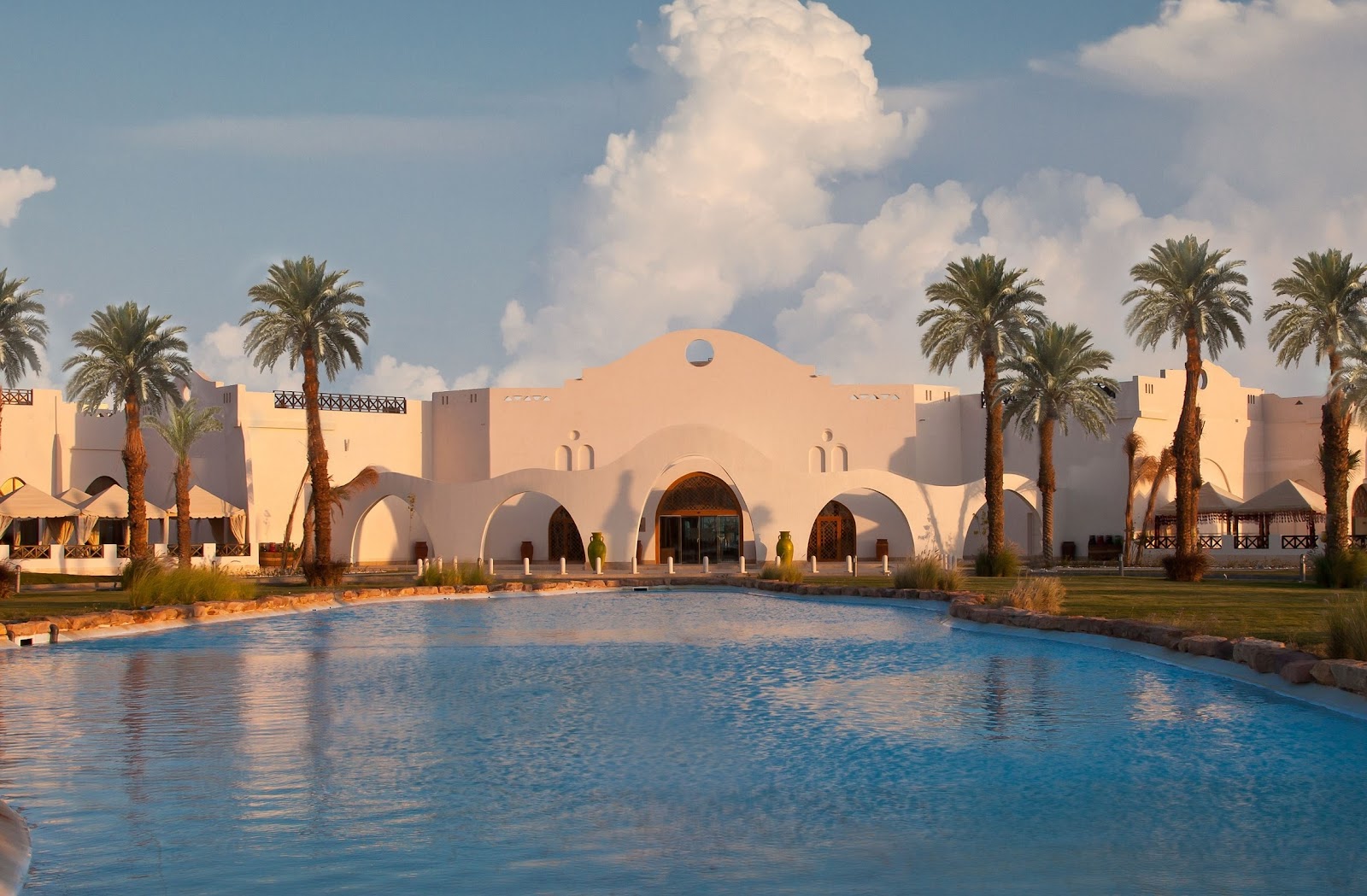 Beige-walled resort building with arched entrances and domed roof overlooks blue pool lined with palm trees under cloudy sky.
