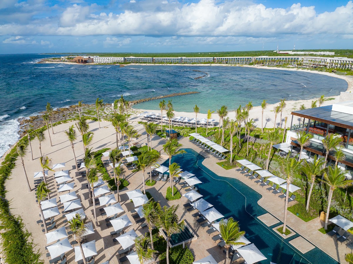Hilton Tulum Riviera Maya resort overlooks a curved beach and turquoise cove, with palm-shaded loungers, a pool, and multi-story hotel buildings along the sandy shore.