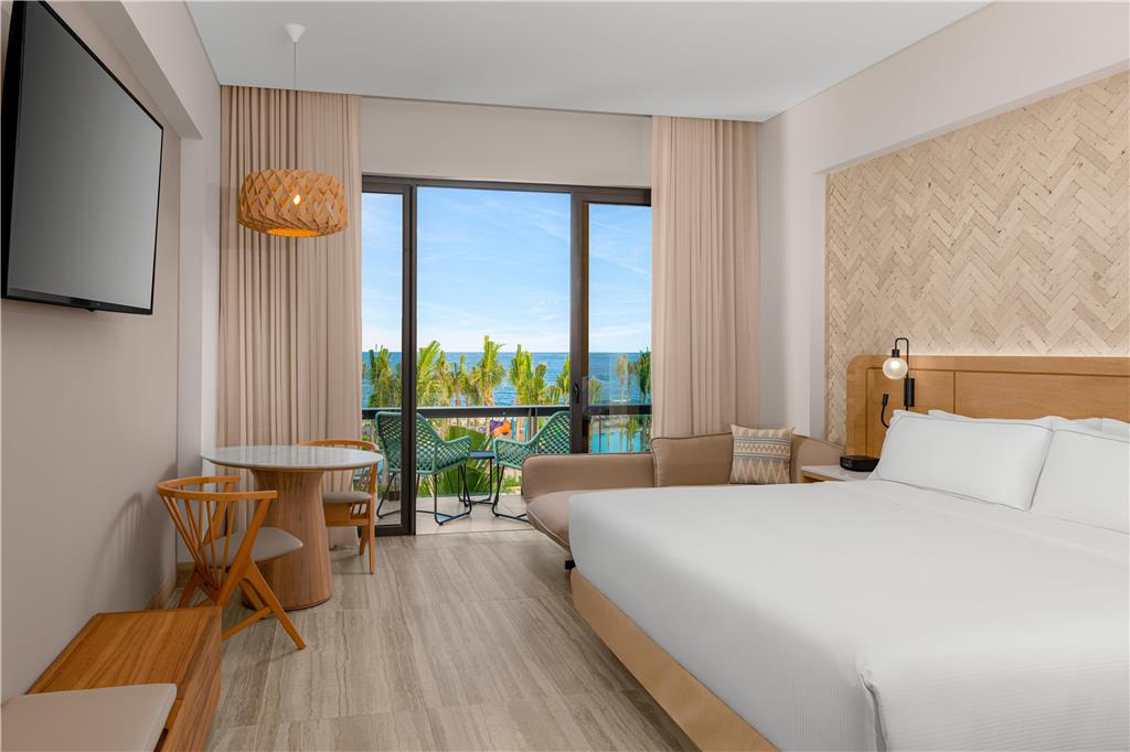Guest room at Hilton Tulum Riviera Maya with queen bed, seating area, and balcony overlooking tropical landscaping and ocean.