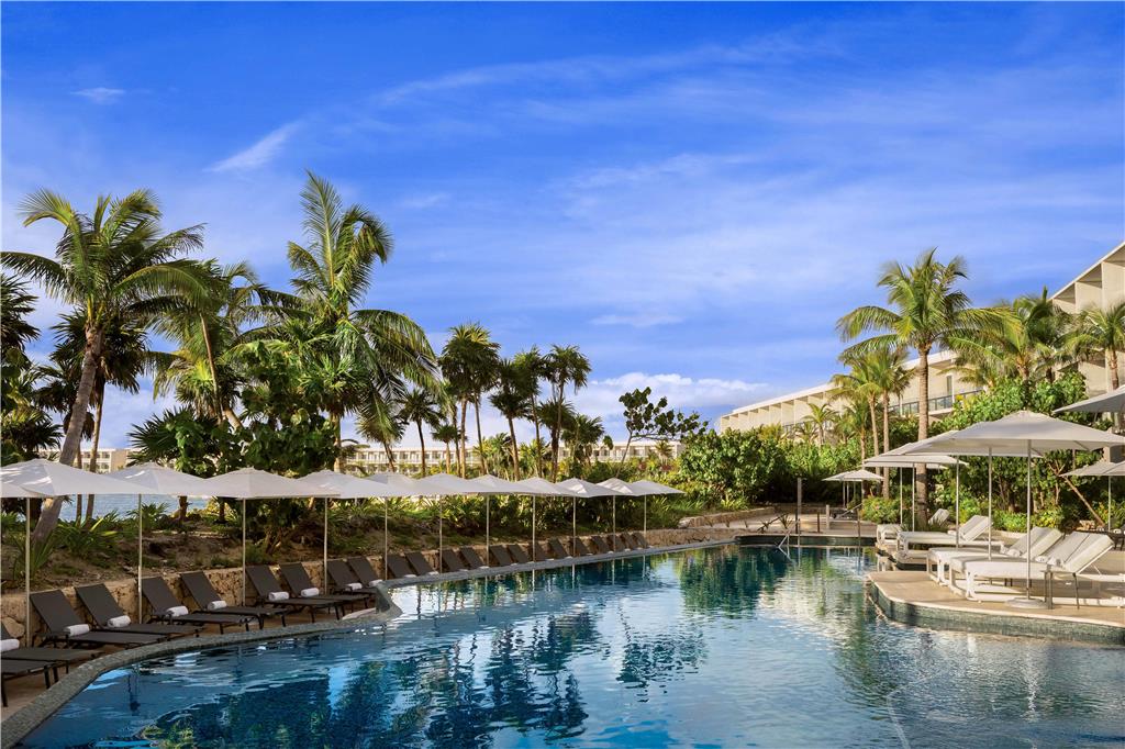Resort pool with loungers and white umbrellas, palm trees, and tan buildings under clear blue sky at Hilton Tulum Riviera Maya.