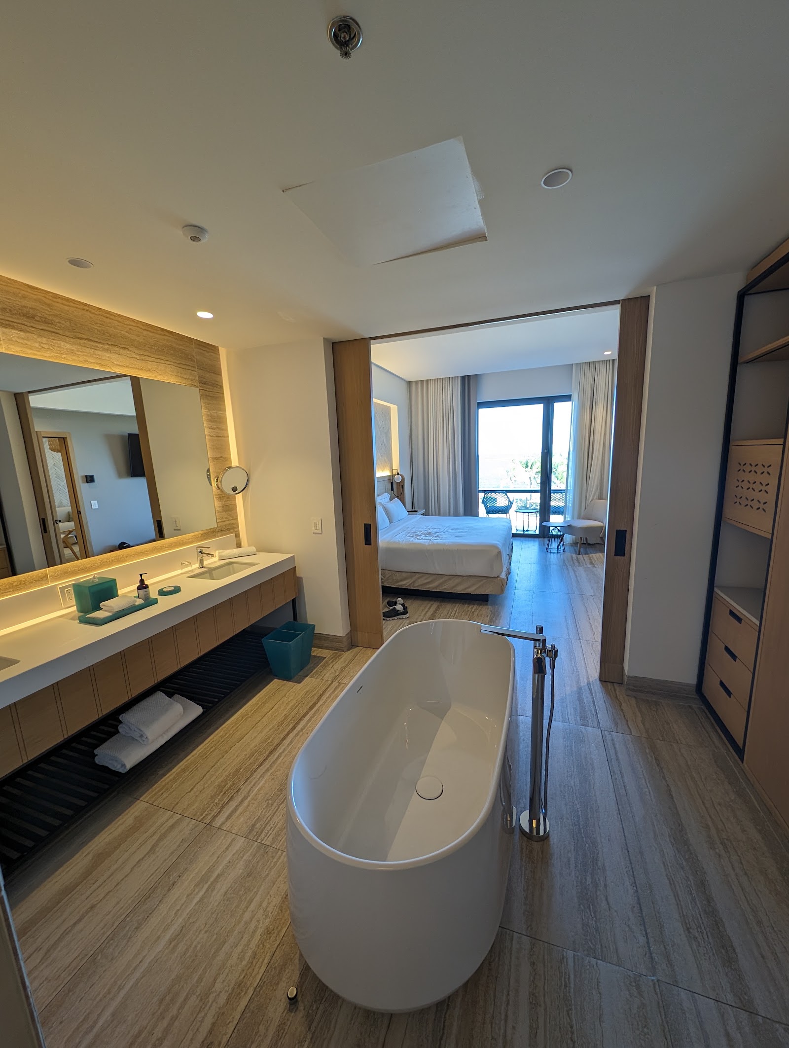 Modern resort bathroom with freestanding white tub, wood vanity, and open doorway to bedroom with ocean view balcony.
