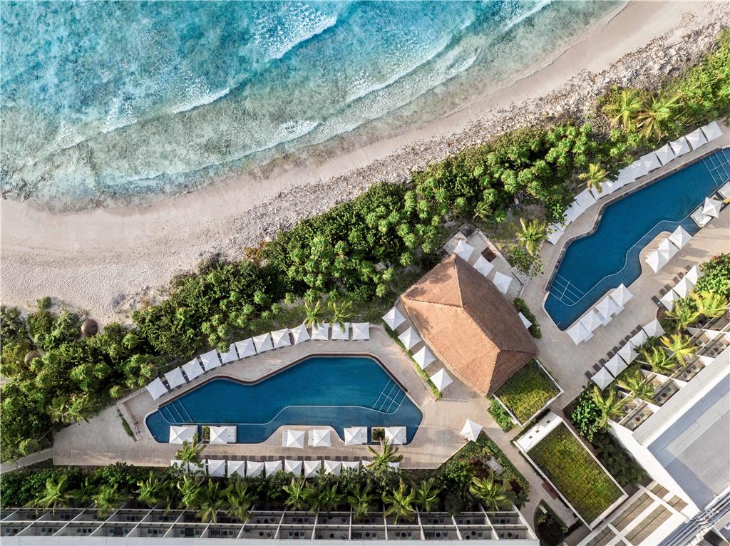 Aerial view of Hilton Tulum Riviera Maya with two curved pools, white loungers, thatched-roof structure, palm trees, and turquoise ocean waves meeting sandy beach.