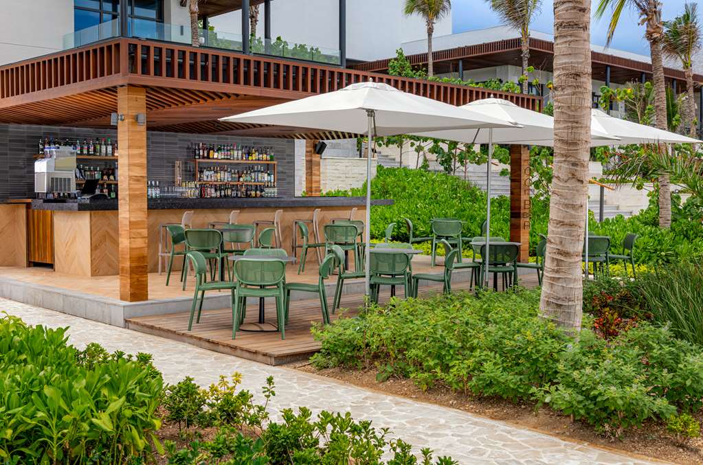Hilton Tulum Riviera Maya's wooden bar pavilion with green chairs under white umbrellas, surrounded by palm trees and tropical landscaping.
