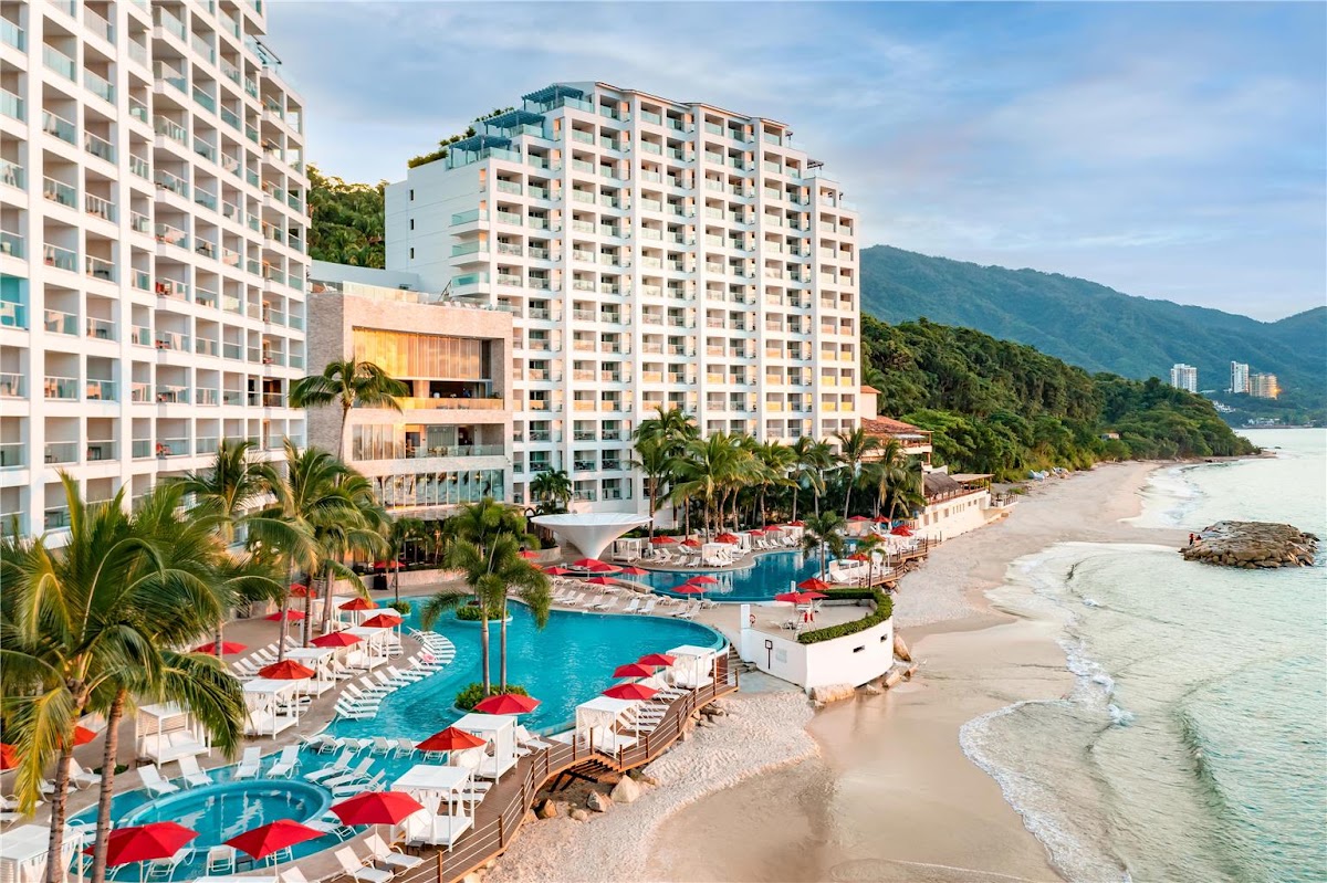 Hilton Vallarta Riviera beachfront with multi-story hotel towers, blue pools with red umbrellas, palm trees, and sandy beach with ocean waves and forested mountains beyond.
