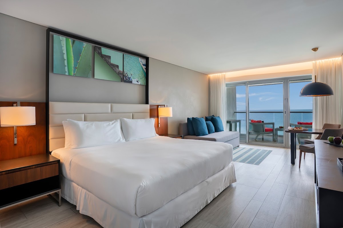 Modern resort bedroom with king bed, ocean view balcony, and coastal scenery visible through floor-to-ceiling glass doors.