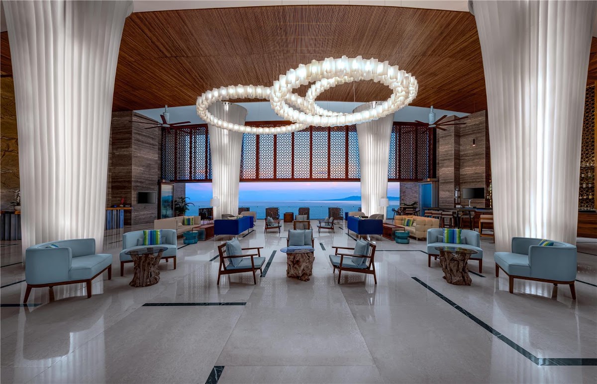 Modern resort lobby with sculptural white light fixture, blue seating areas, and ocean view through large windows at Hilton Vallarta Riviera.