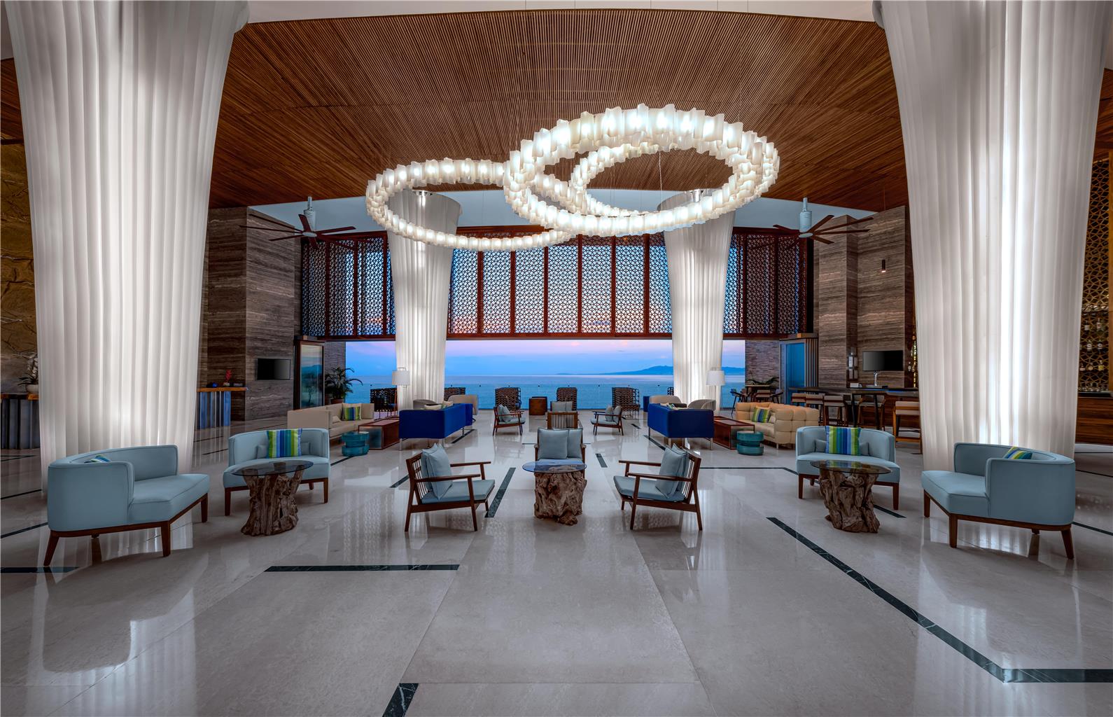 Red lounge chairs with a bowl of apples on a balcony overlooking a sandy beach, pool, and turquoise ocean with a white resort building and mountain beyond.