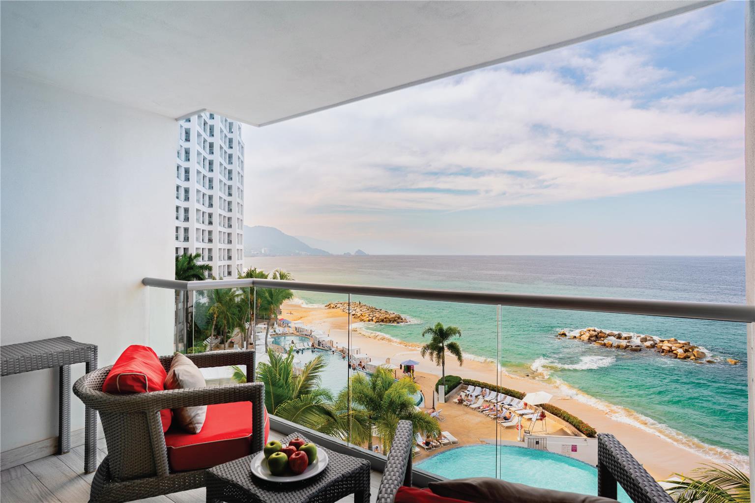Modern restaurant dining room with wooden tables and red cushioned chairs, white pendant lights overhead, and ocean view through floor-to-ceiling windows at Hilton Vallarta Riviera.