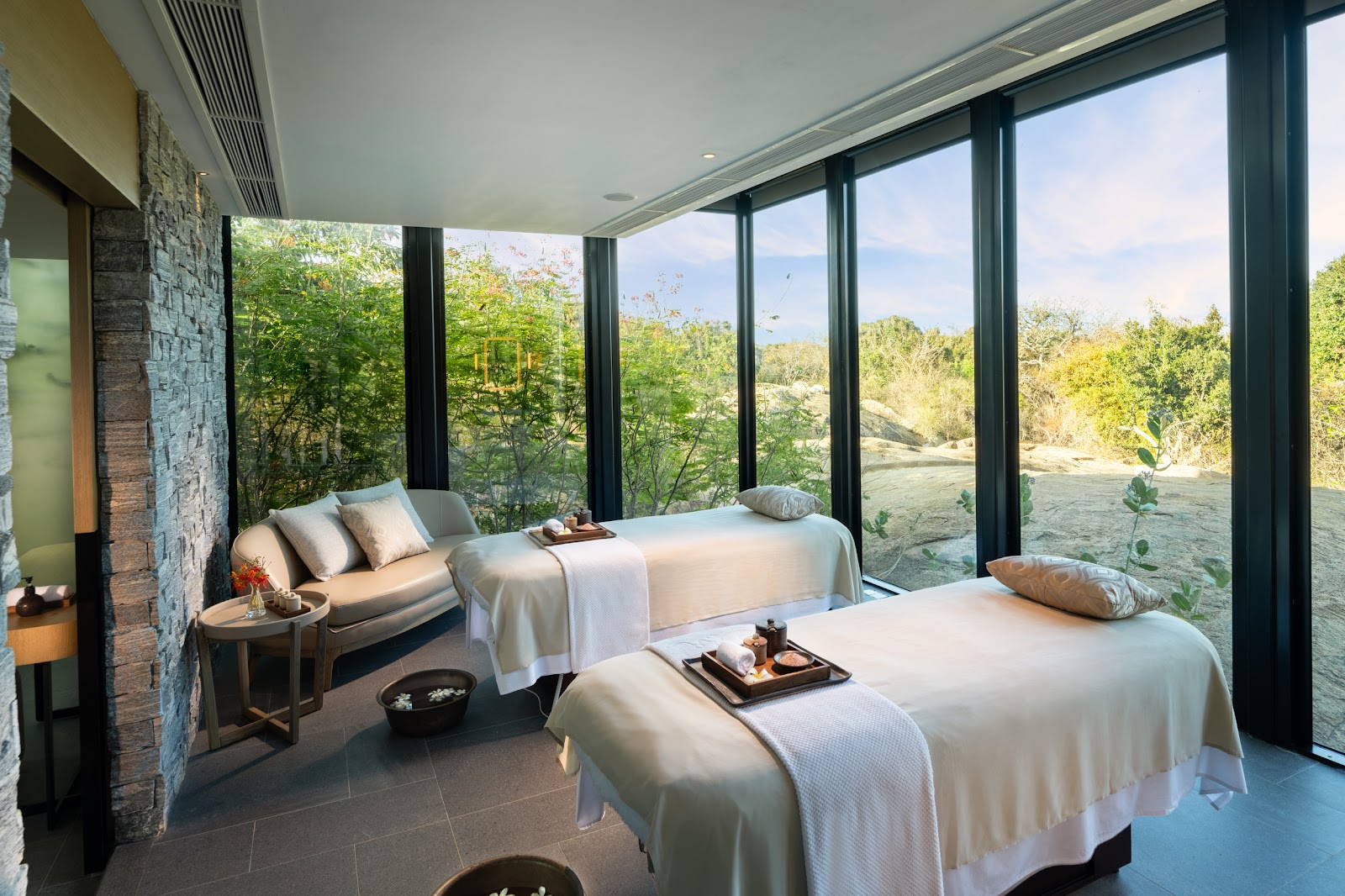 Spa treatment room with two massage beds, stone wall, and floor-to-ceiling windows overlooking forested hillside at Hilton Yala Resort.