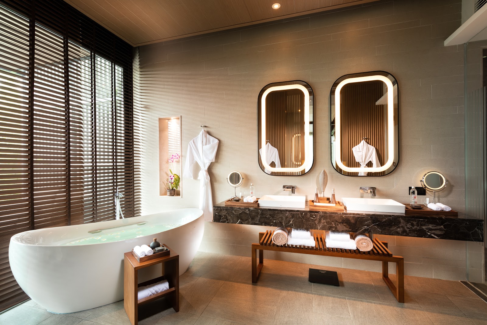 Spacious bathroom with dual vanity, illuminated mirrors, walk-in shower, and freestanding white bathtub with slatted window shutters.
