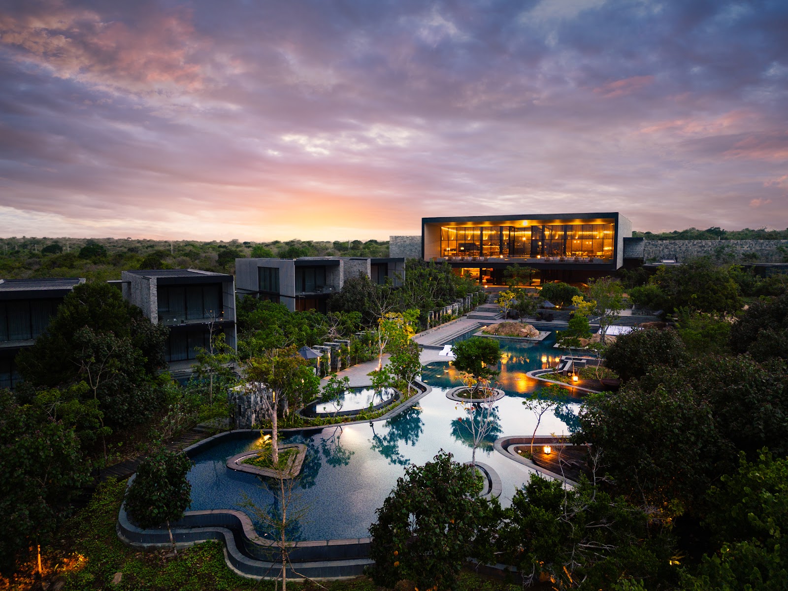 Hilton Yala Resort at dusk with illuminated modern pavilions, curved pools, and jungle vegetation under a purple and pink sky.
