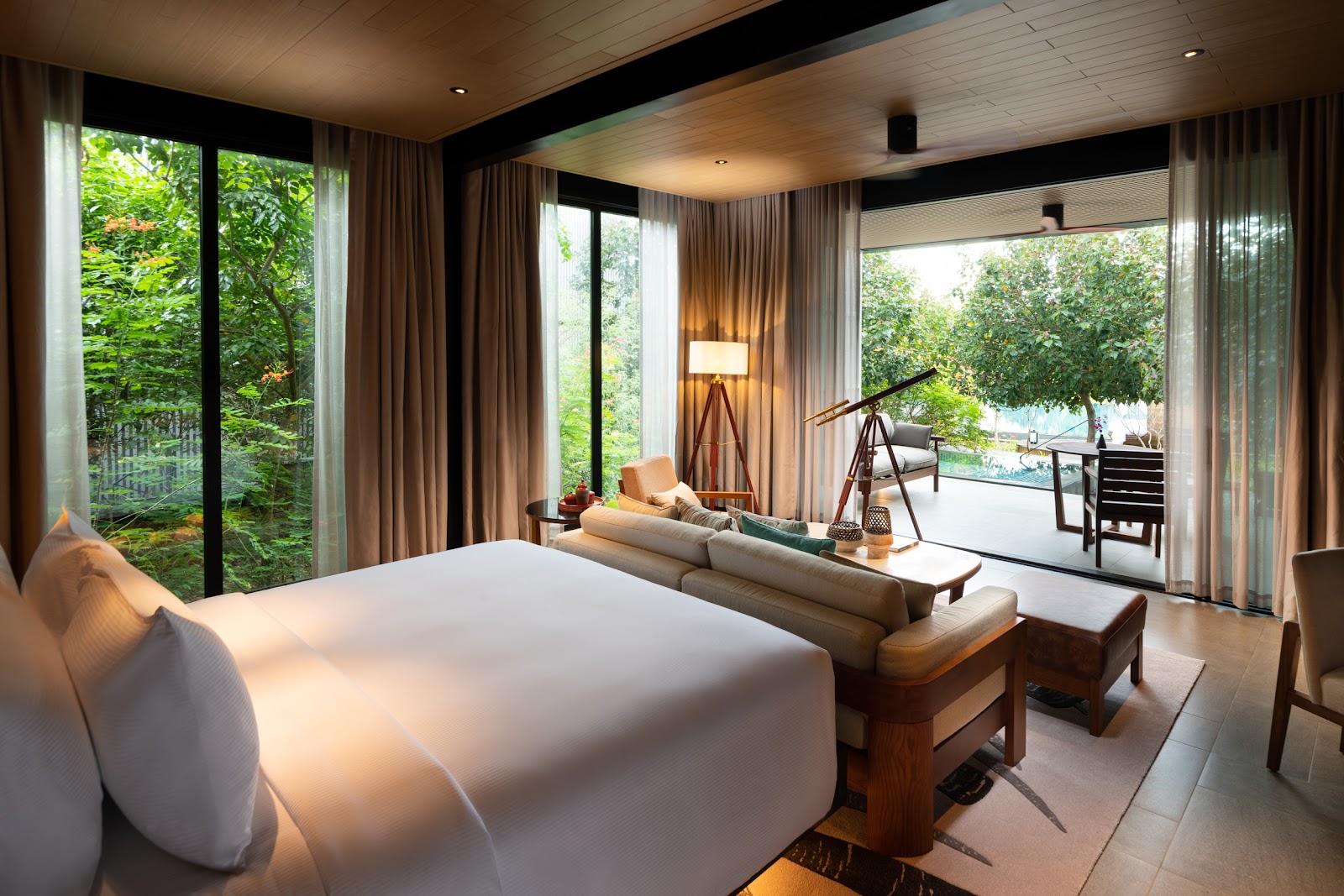 Spacious resort villa with white bed, living area, and floor-to-ceiling windows overlooking lush tropical forest and pool beyond.