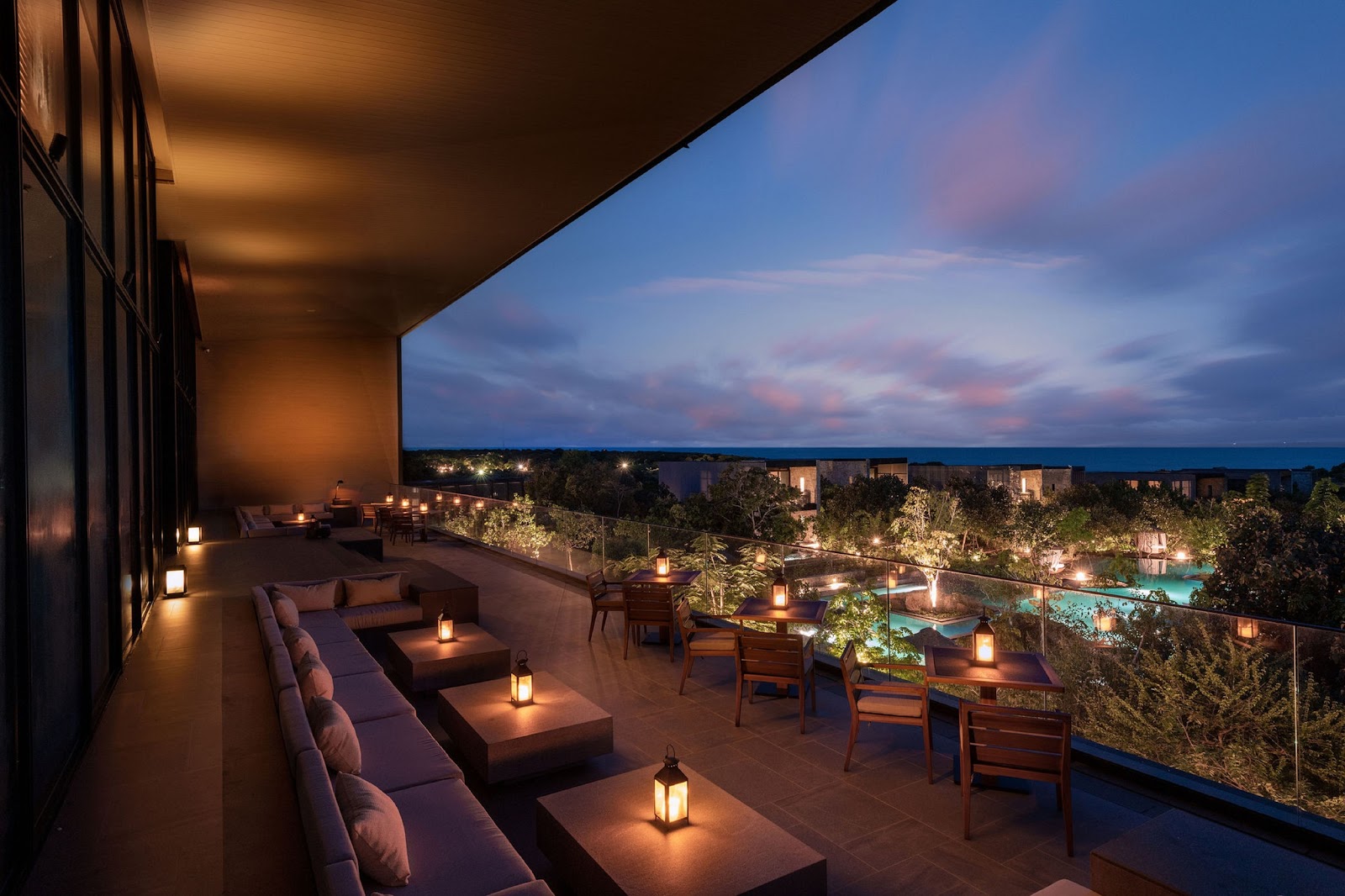 Elevated terrace at dusk with lounge seating, lanterns, and views of illuminated grounds, pool, and ocean beyond.