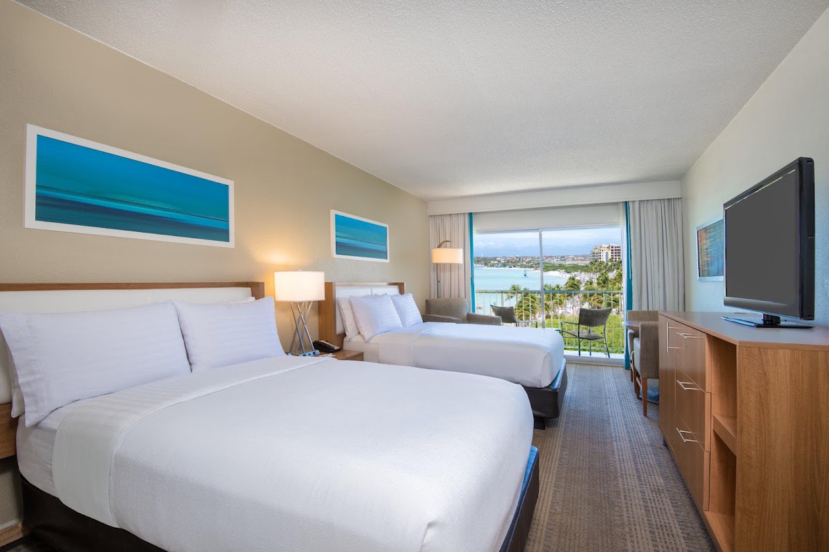 Resort room with two queen beds, flat-screen TV, and a balcony overlooking Aruba's turquoise bay and beachfront.