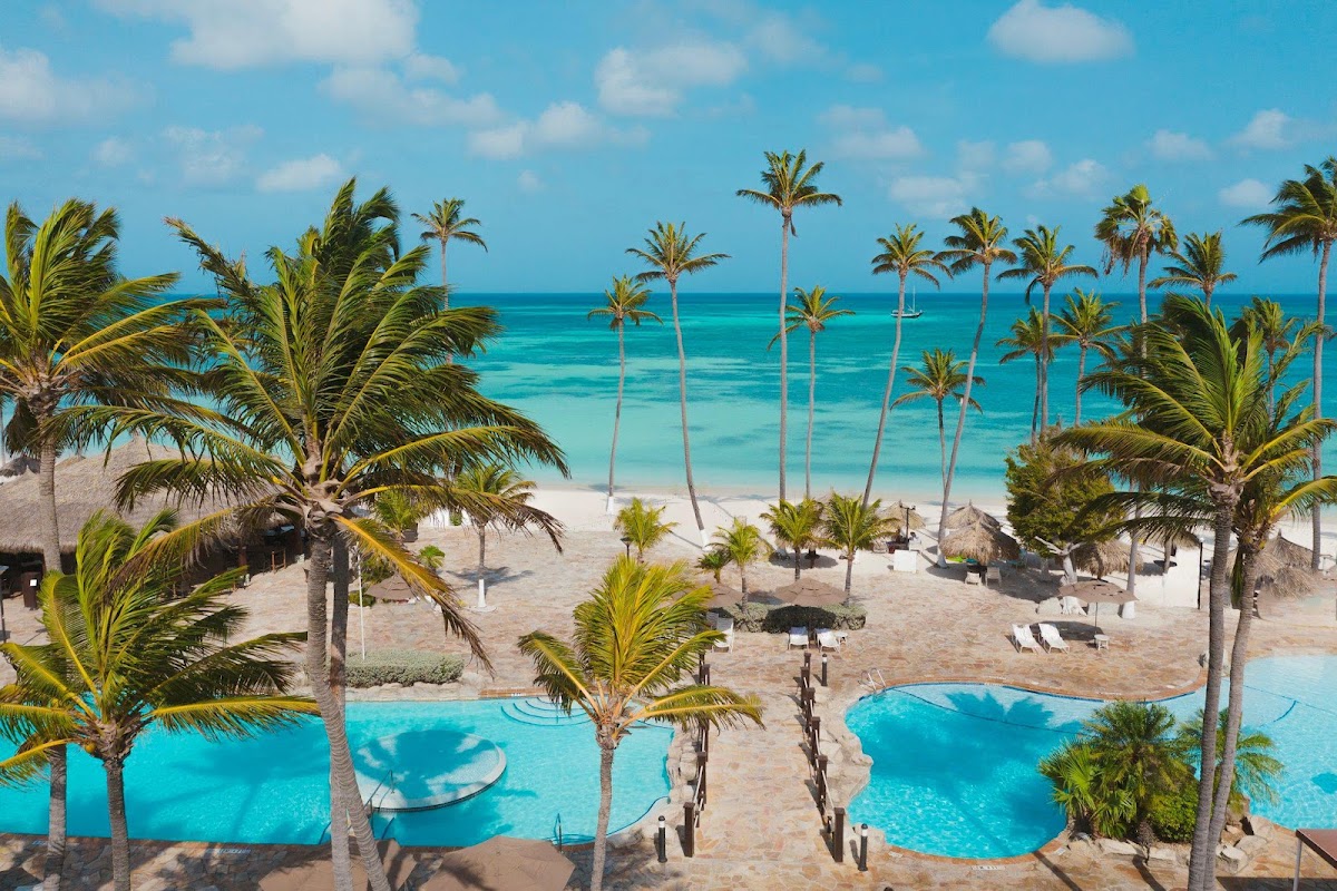 Aerial view of Holiday Inn Resort Aruba with curved pools, palm trees, beach, and turquoise ocean with sailboat.