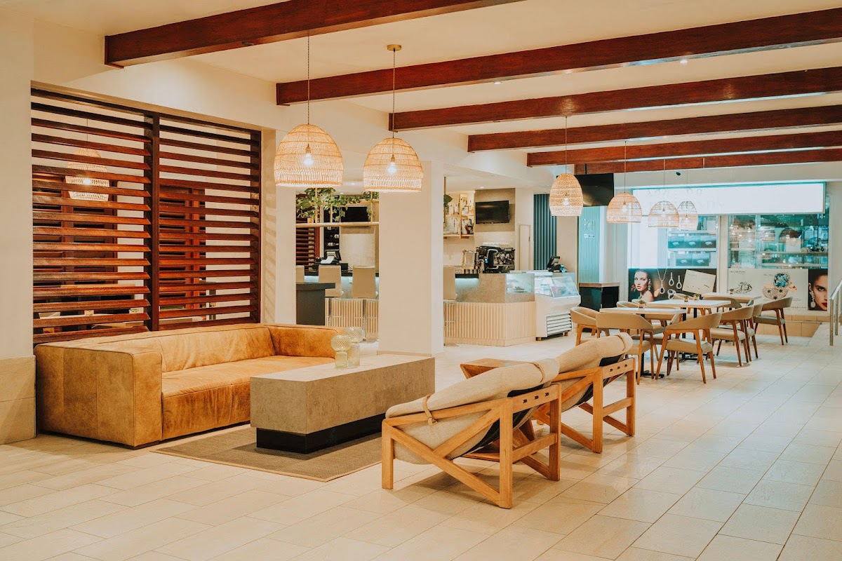 Resort lobby with tan tile floors, cognac leather sofa, wooden beam ceiling, woven pendant lights, and dining area visible in background.