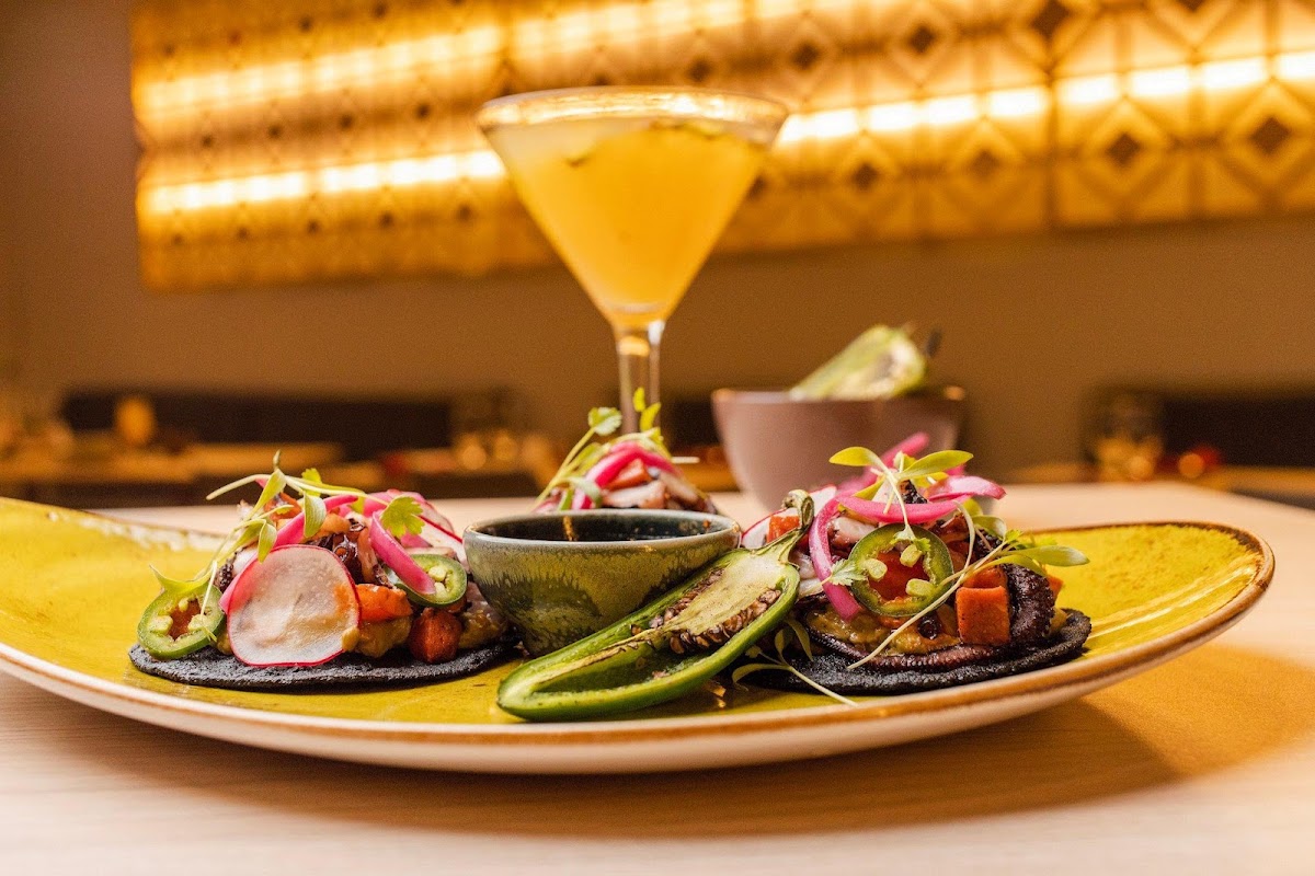 Plated appetizer with black tortillas, radish slices, and microgreens beside a yellow cocktail under warm resort lighting.