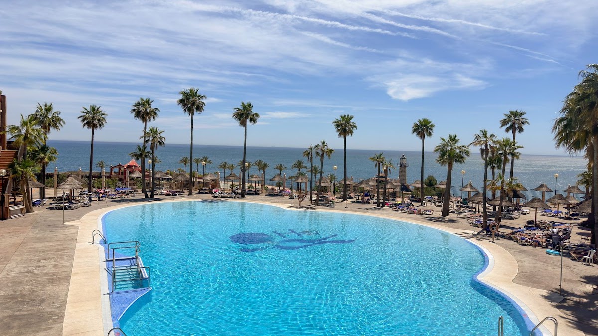 Large curved pool with blue water overlooking the Mediterranean Sea, palm trees, and beach loungers at Holiday World Polynesia.