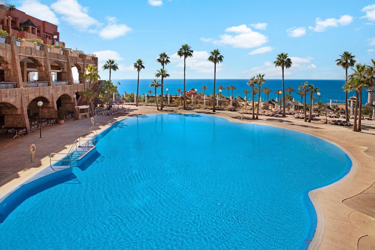 Large curved pool with terracotta resort buildings, palm trees, and Mediterranean Sea beyond under blue sky.