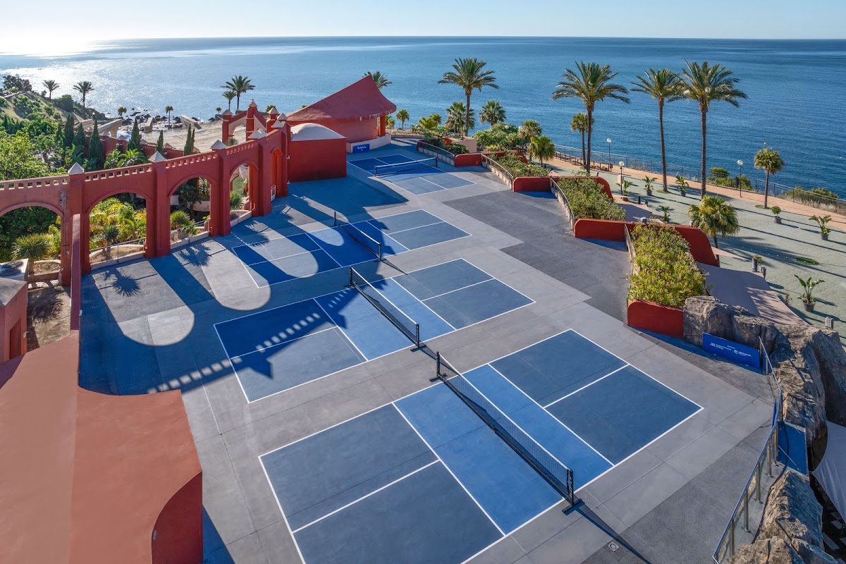 Tennis and padel courts at Holiday World Polynesia overlook the Mediterranean Sea, framed by red architectural elements and palm trees.