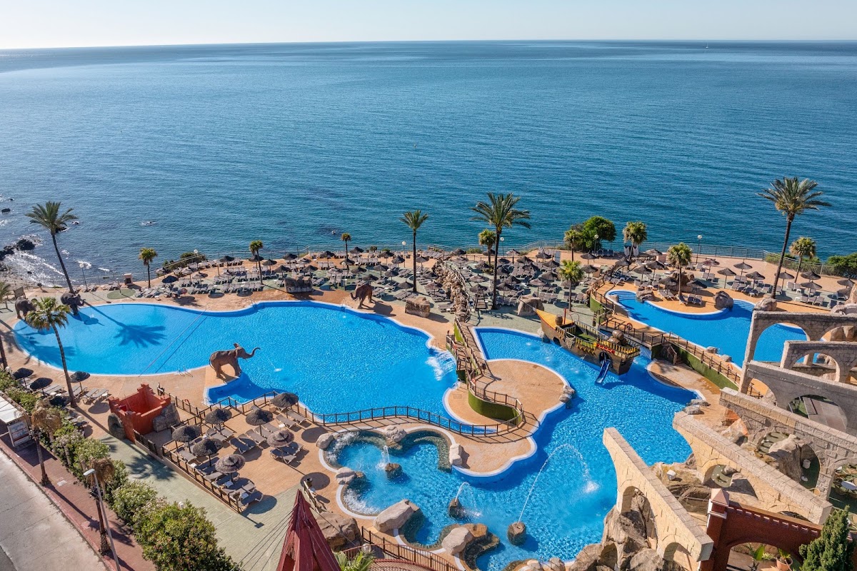 Aerial view of Holiday World Riwo pool complex with multiple blue pools, palm trees, beach lounge chairs, and Mediterranean Sea beyond.
