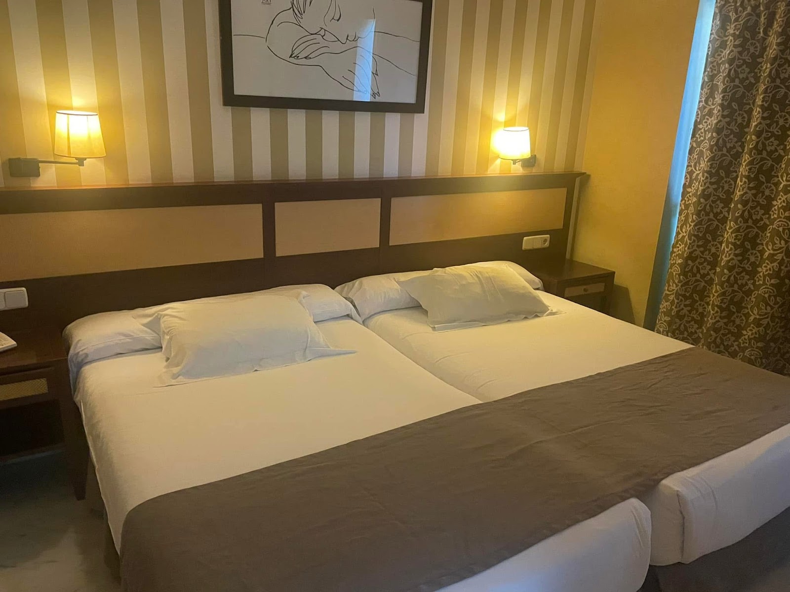 Hotel room with two beds with white pillows and linens, striped gold and white walls, and a framed line drawing above the headboard.