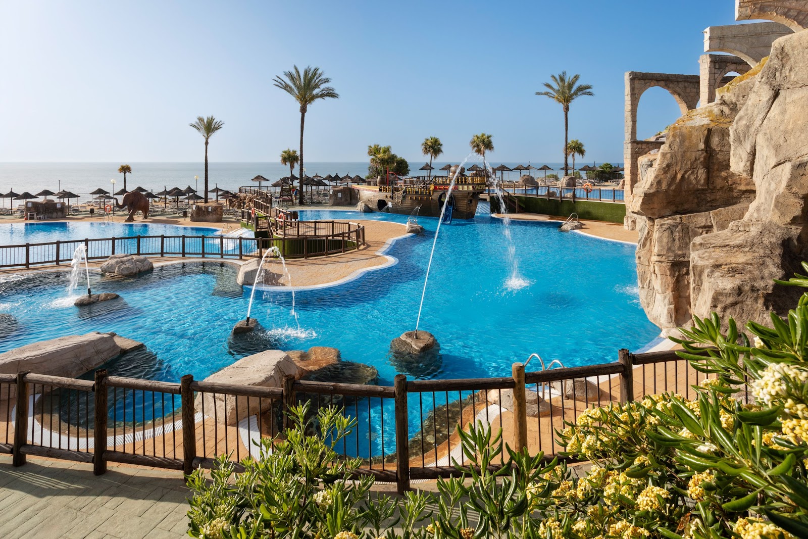 Multiple turquoise pools with fountains, palm trees, and beach umbrellas at Holiday World Riwo, with the Mediterranean Sea visible beyond.