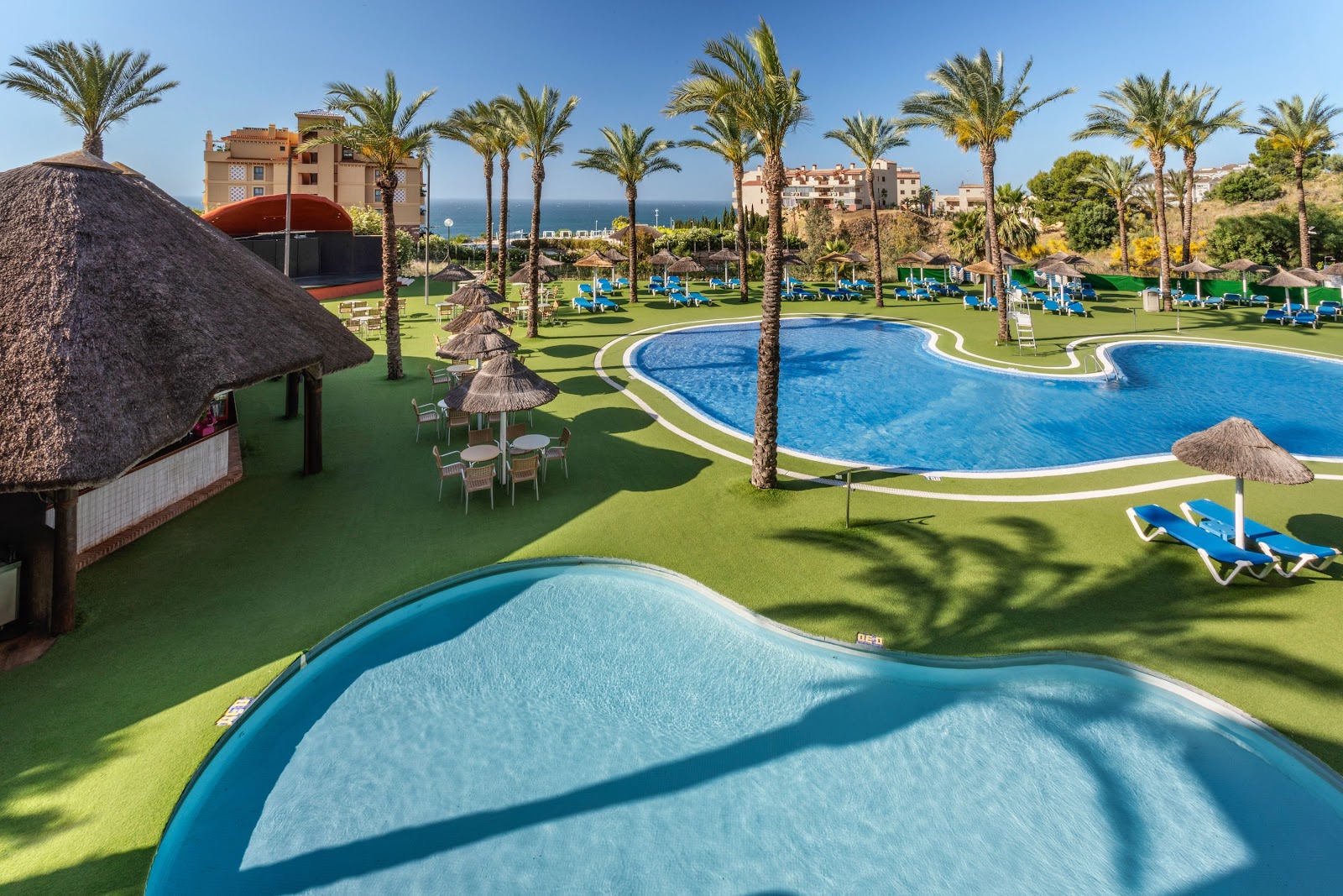 Resort pools with blue loungers and thatched umbrellas on manicured lawns, overlooking the Mediterranean Sea and resort buildings.