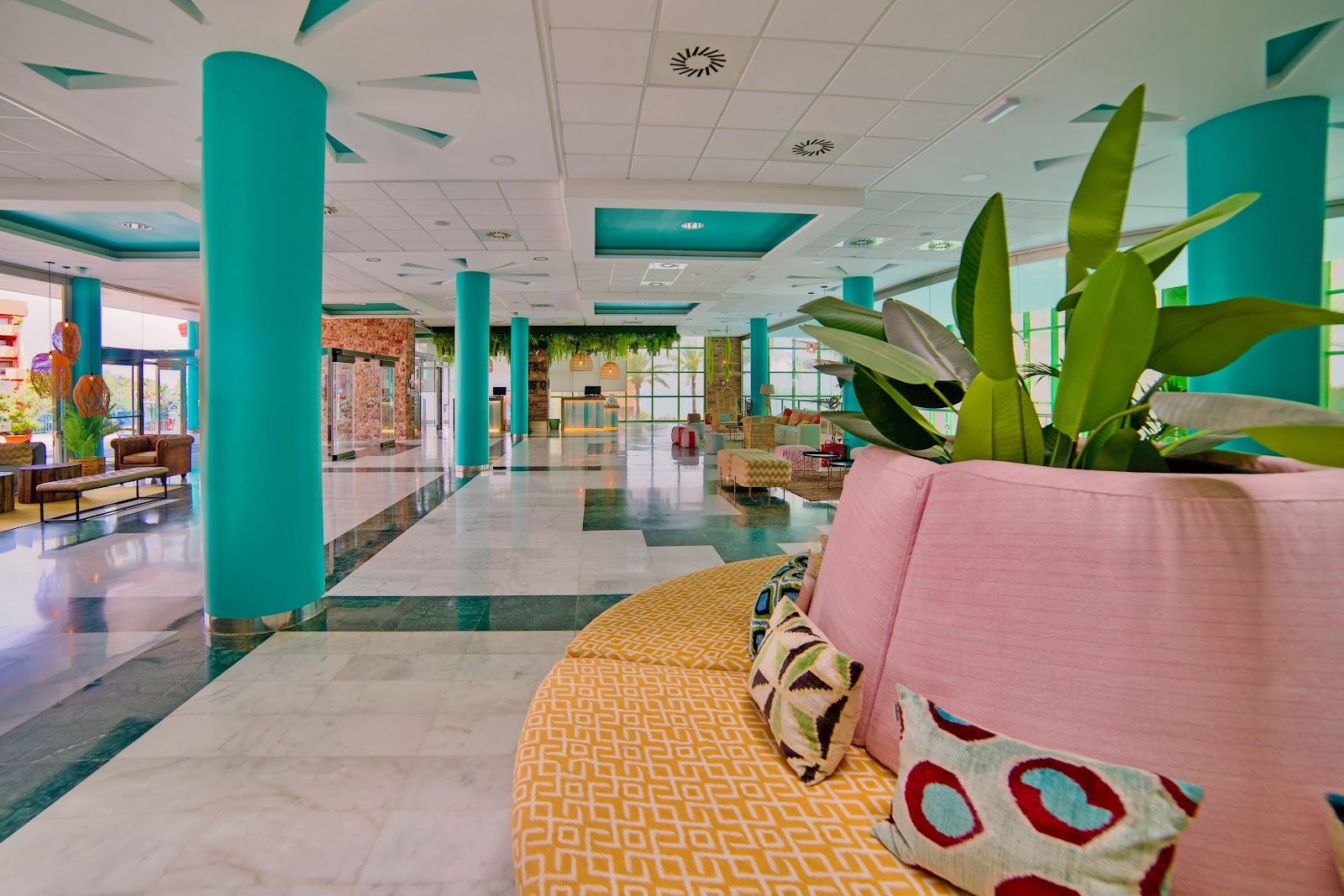 Bright modern lobby with turquoise columns, green-framed windows, colorful furniture in pink and yellow tones, and hanging wire pendant lights.