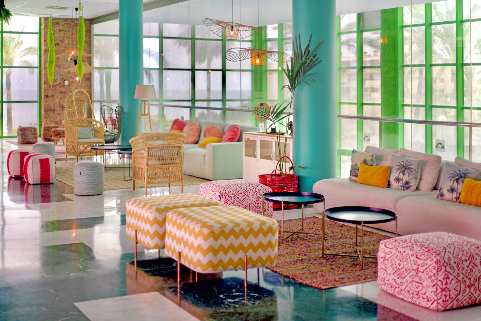 Modern dining area with red and green chairs at wooden tables, tall yellow columns, and tropical plants under a multi-story atrium at Holiday World Riwo.