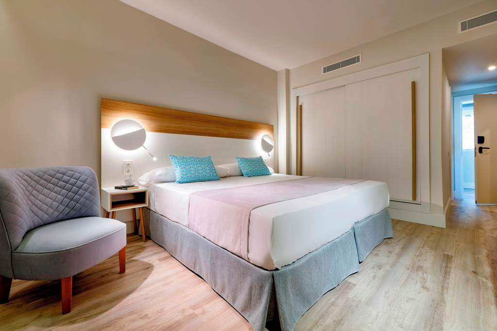 Modern hotel room with queen bed, turquoise pillows, wooden headboard with round sconces, gray accent chair, and light wood flooring.