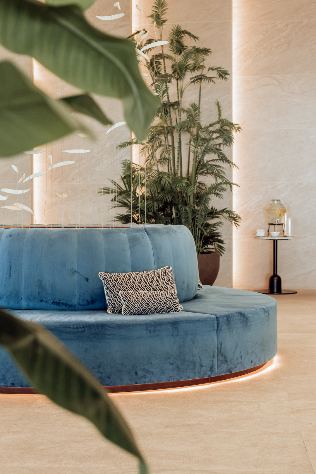 Blue curved seating with geometric pillow in a modern lobby with potted palms, beige walls, and natural light at Hotel Benalma Costa del Sol.