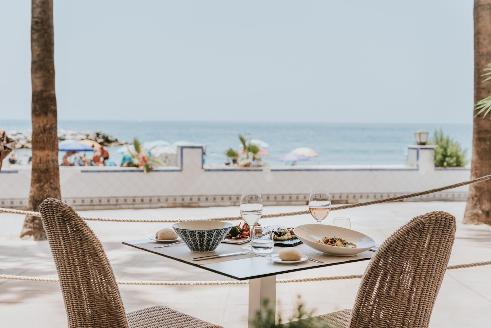 Two woven chairs at a waterfront table with plated food and wine glasses, ocean and beach beyond.