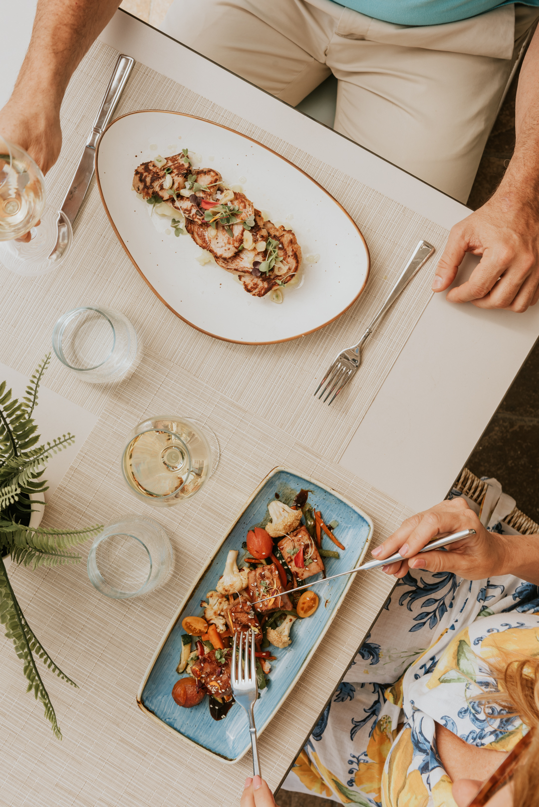 Two diners enjoy plated entrees at a beige table, with grilled meat on white oval plate and roasted vegetables on blue rectangular plate, white wine glasses visible.
