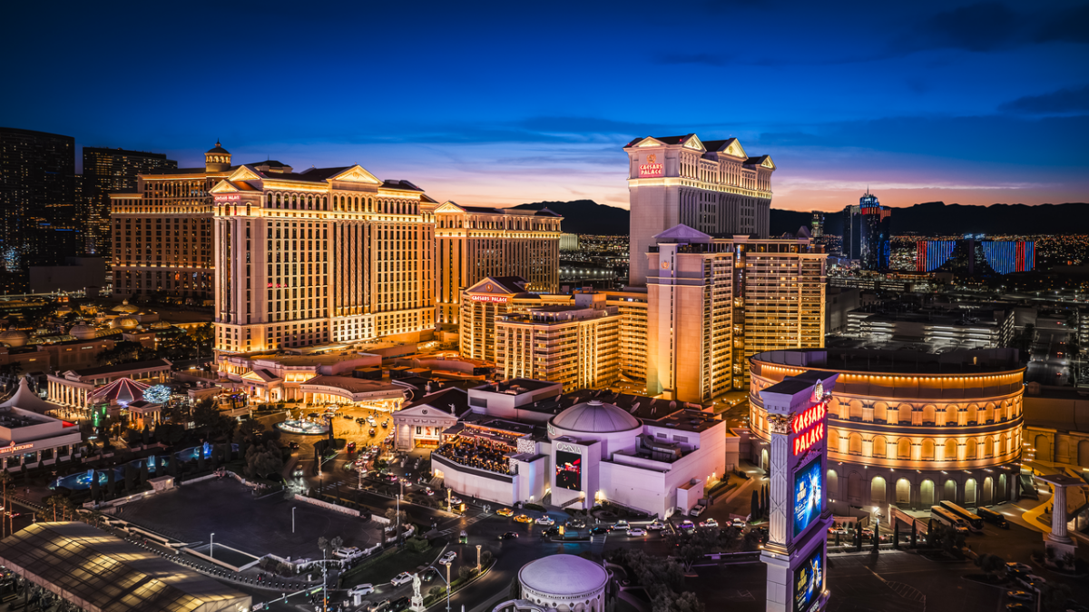 Illuminated resort buildings and casino structures line a Las Vegas strip at twilight, with mountains visible in the distant background.