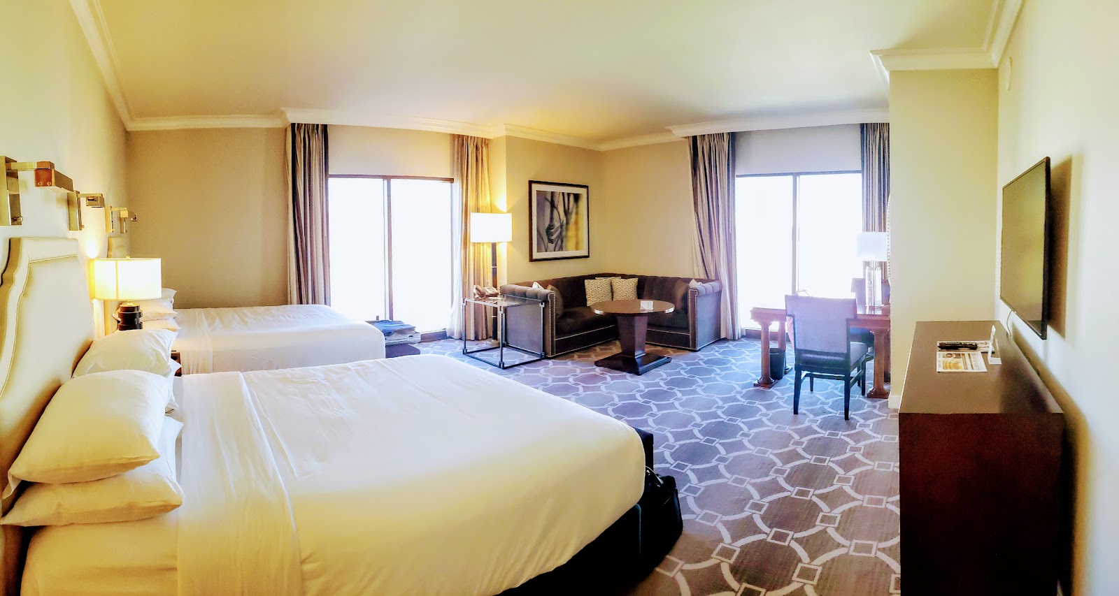 Spacious resort room with two queen beds, seating area with dark sofa, patterned carpet, and balcony doors letting in natural light.