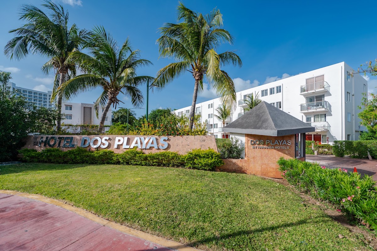 Hotel Dos Playas entrance with signage, palm trees, manicured lawn, and white multi-story resort buildings under clear blue sky.