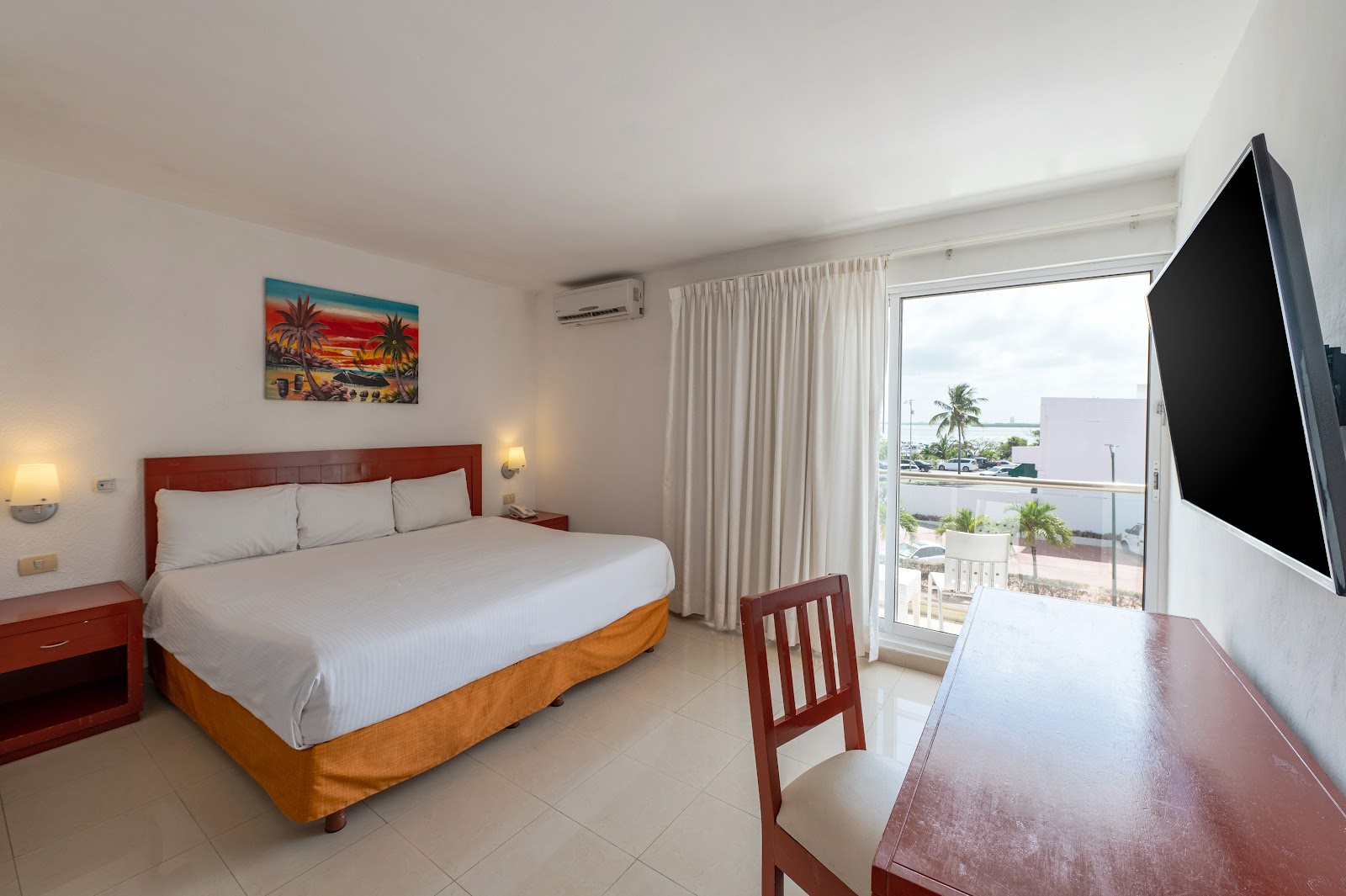 Hotel room with queen bed, red headboard, white bedding, and balcony overlooking palm trees and ocean at Hotel Dos Playas Cancún.