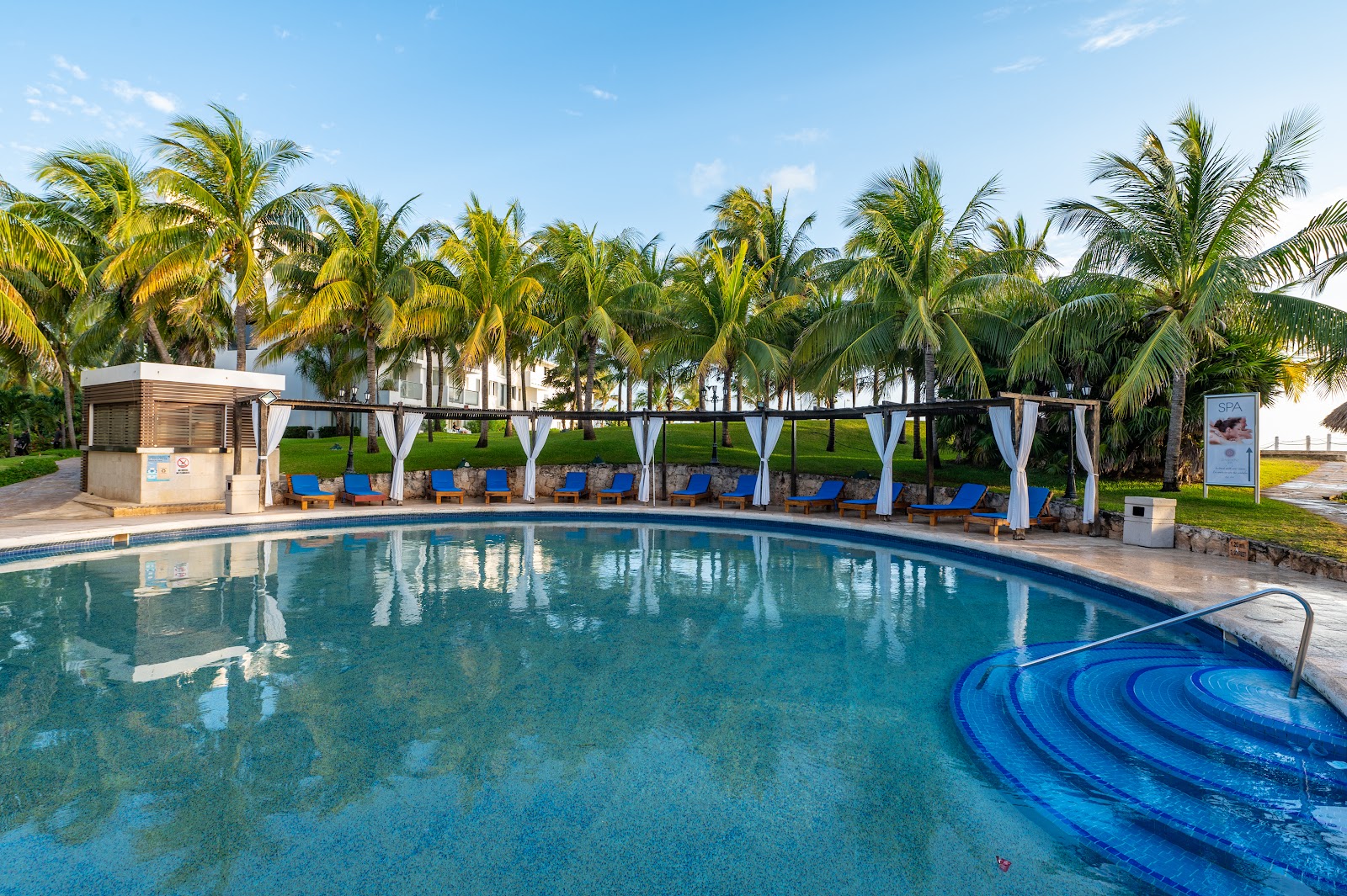 Curved pool with blue loungers under white pergolas and palm trees at Hotel Dos Playas Cancún.