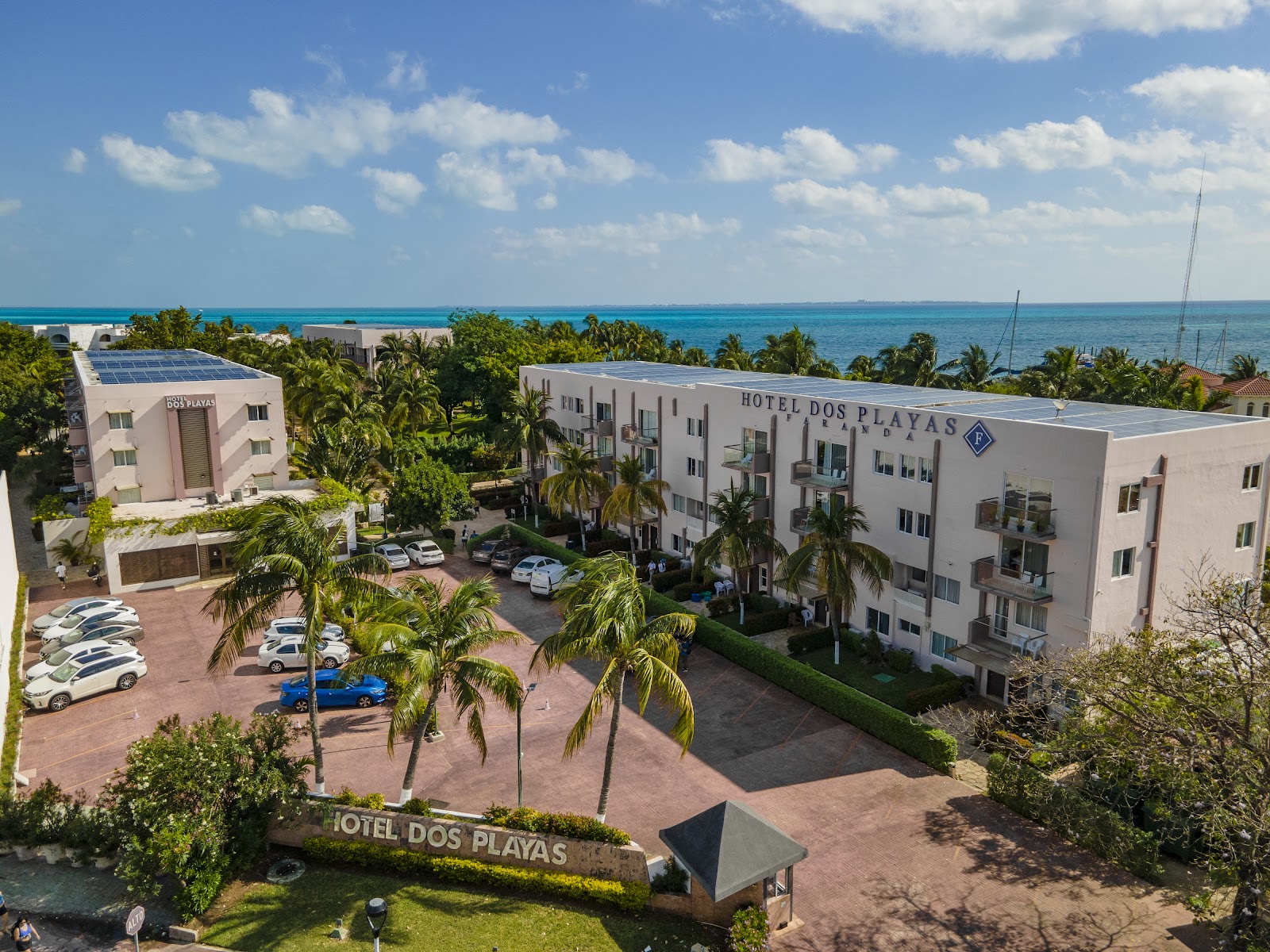 Hotel Dos Playas Cancún's white multi-story buildings overlook a palm-lined courtyard with parked cars and the turquoise Caribbean Sea beyond.