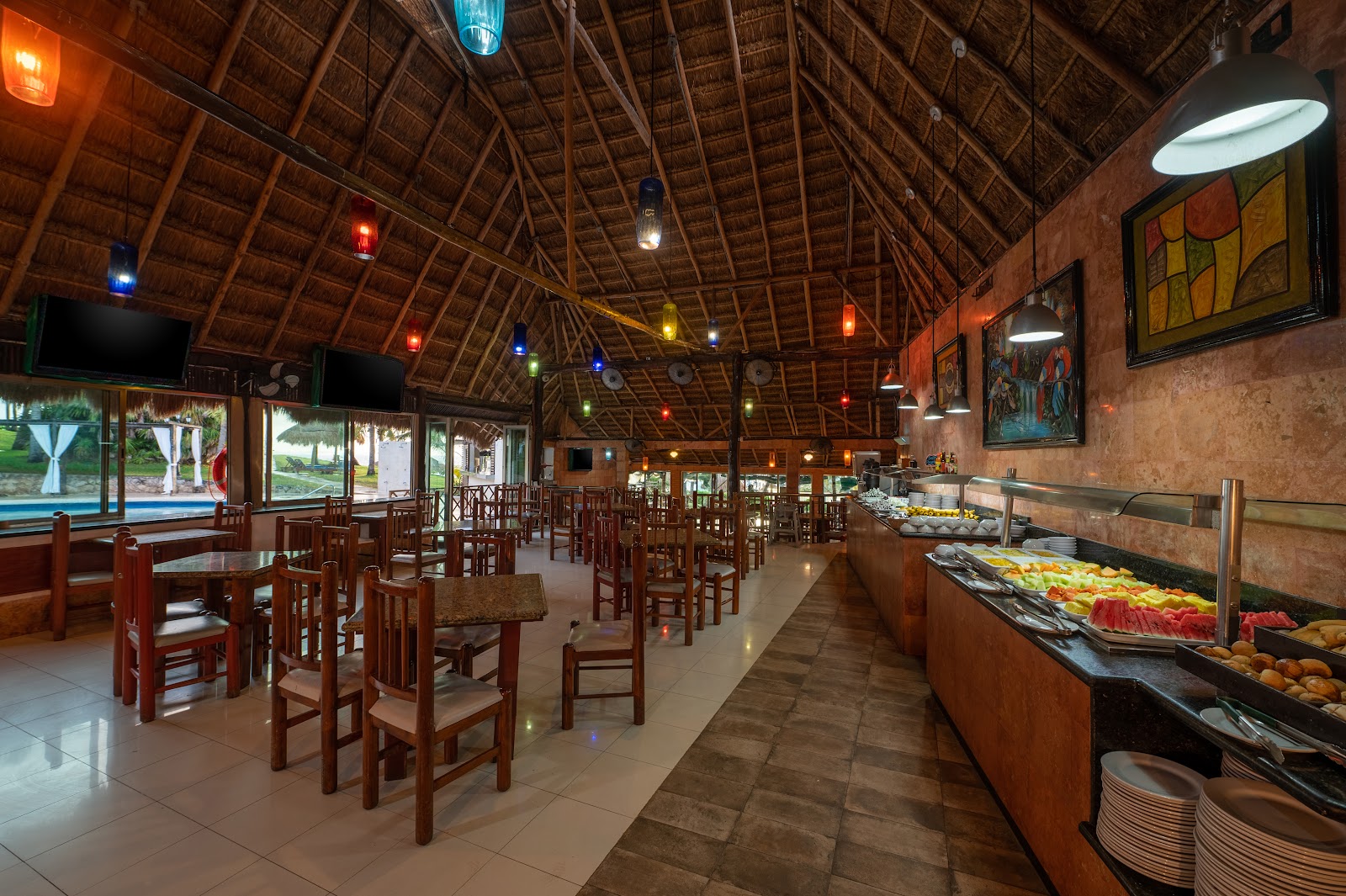 Dining hall with wooden chairs and tables, buffet stations displaying fresh fruits and vegetables, thatch roof with colorful pendant lights, and views of the pool area through windows.