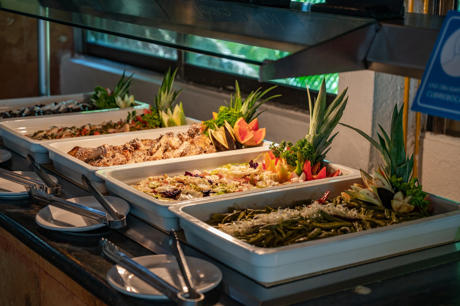 White serving trays of salads and grilled vegetables garnished with colorful flowers and tropical plants at a buffet station.