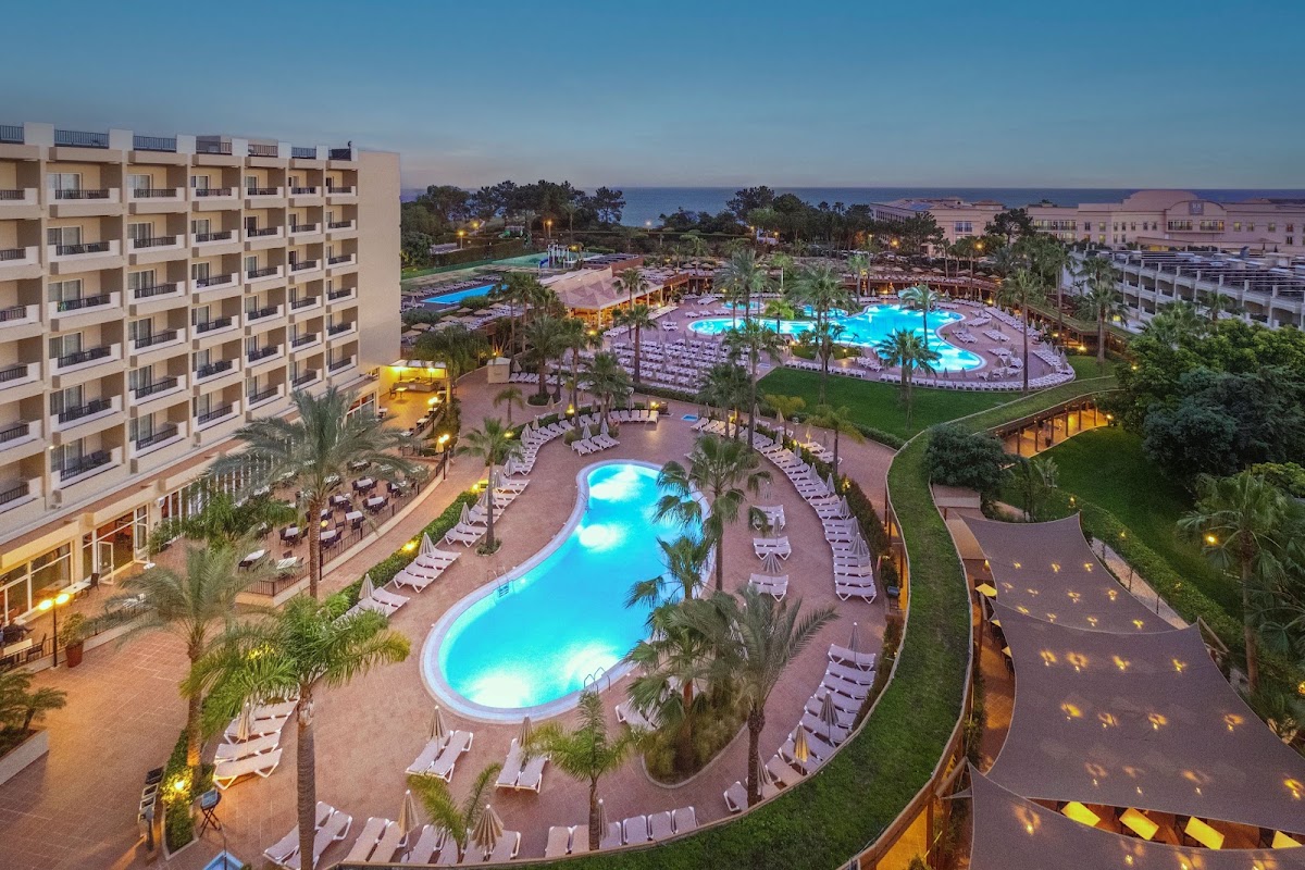 Resort grounds at dusk with multiple illuminated pools, loungers, palm trees, and beige hotel buildings overlooking the sea beyond.