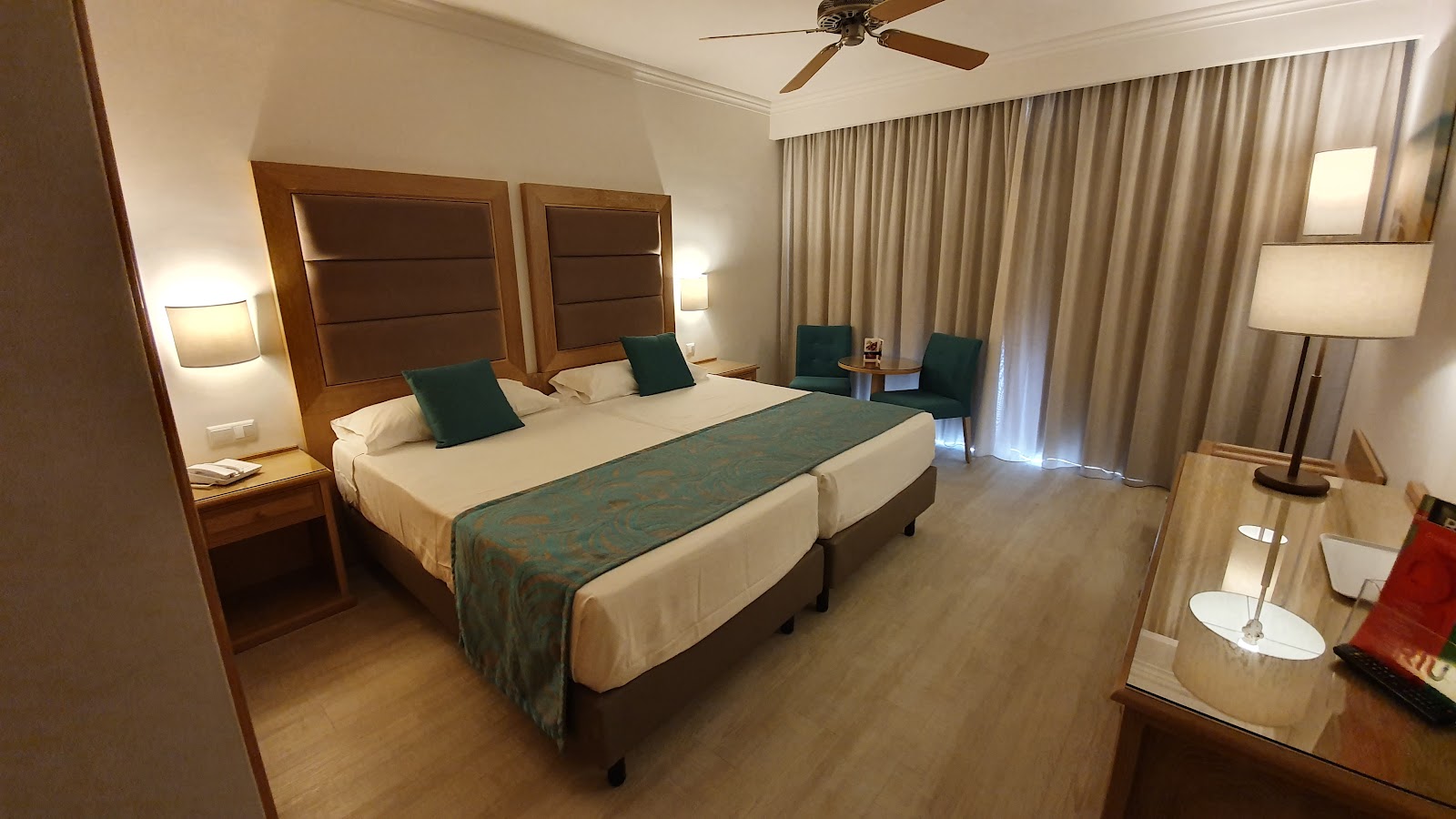 Hotel Guarana guest room with two beds, green accent pillows, wooden headboards, bedside lamps, ceiling fan, and floor-to-ceiling curtains.